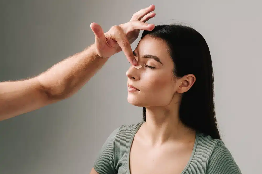 Surgeon examining young woman’s nose for signs of asymmetry before potential revision rhinoplasty consultation.