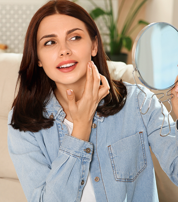 Woman examining her face in a mirror, considering non-surgical facial enhancement alternatives for chin and jawline.